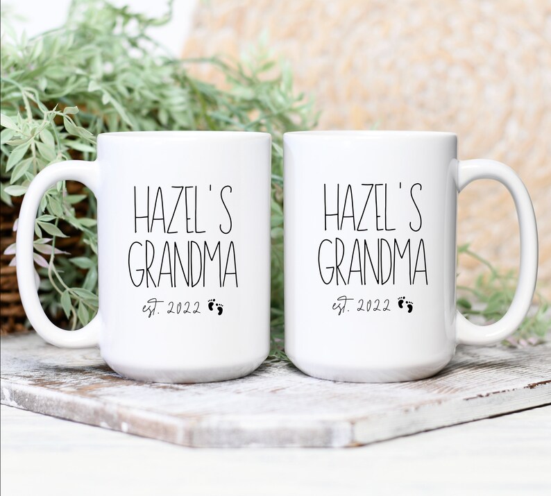 Op de afbeelding: Twee witte keramische mokken met zwarte tekst die "Hazel's Grandma est. 2022" met een paar voetafdrukken zegt.