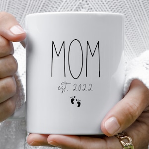 May include: White ceramic mug with black text that reads "MOM est. 2022" with two small black footprints below the text.