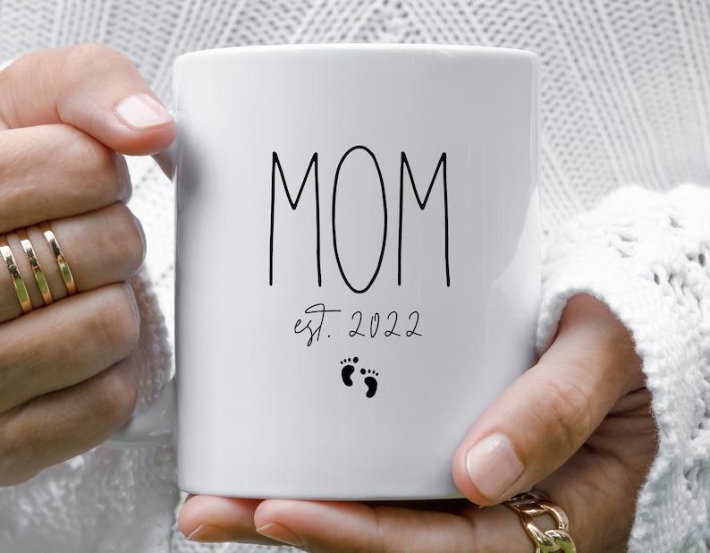 May include: White ceramic mug with black text that reads "MOM est. 2022" with two small black footprints below the text.