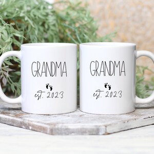 May include: Two white ceramic mugs with black text that reads "GRANDMA" above a pair of baby footprints and "est. 2023". The mugs are on a wooden surface with greenery in the background.