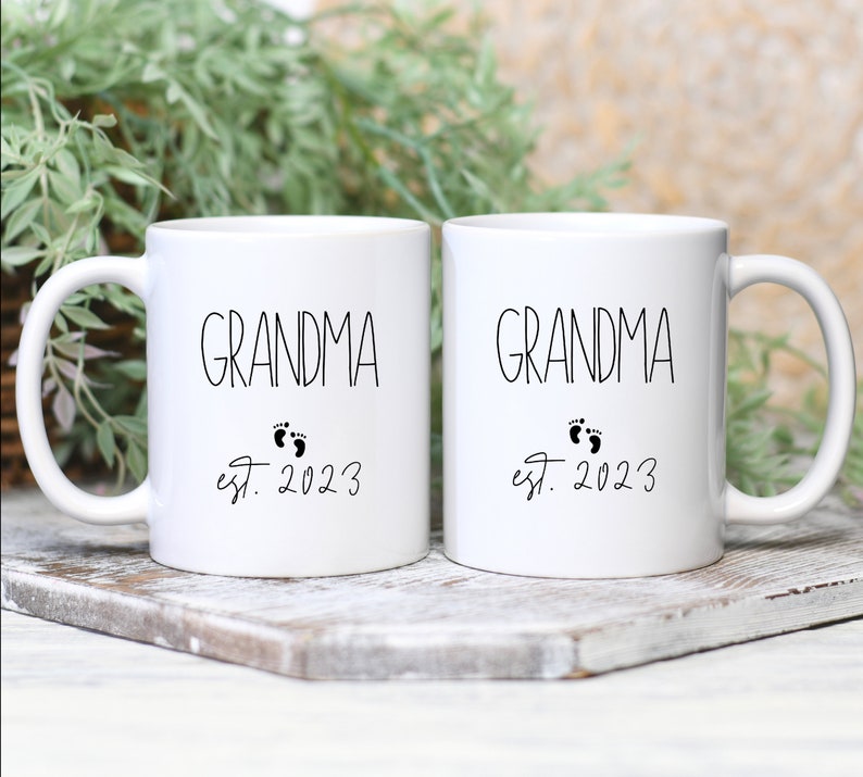 May include: Two white ceramic mugs with black text that reads "GRANDMA" above a pair of baby footprints and "est. 2023". The mugs are on a wooden surface with greenery in the background.