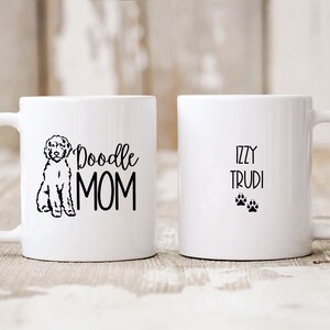 Op de afbeelding: Witte keramische koffiemok met een zwarte doodle-hond-illustratie en de tekst "Doodle Mom". De andere kant van de mok heeft de tekst "Izzy Trudi" en twee zwarte poot afdrukken.