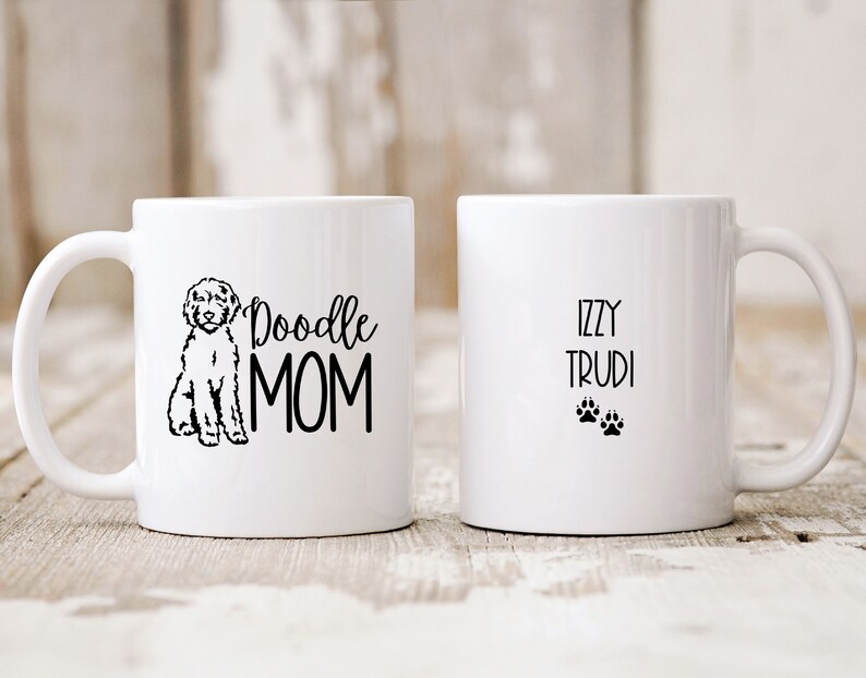 Op de afbeelding: Witte keramische koffiemok met een zwarte doodle-hond-illustratie en de tekst "Doodle Mom". De andere kant van de mok heeft de tekst "Izzy Trudi" en twee zwarte poot afdrukken.