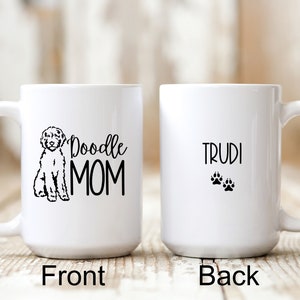 Op de afbeelding: Witte keramische koffiemok met een zwart doodle-hondensilhouet en de tekst "Doodle Mom" aan de voorkant. De achterkant van de mok heeft de tekst "Trudi" en twee zwarte poot afdrukken.