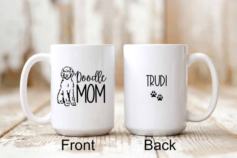 Op de afbeelding: Witte keramische koffiemok met een zwart doodle-hondensilhouet en de tekst "Doodle Mom" aan de voorkant. De achterkant van de mok heeft de tekst "Trudi" en twee zwarte poot afdrukken.