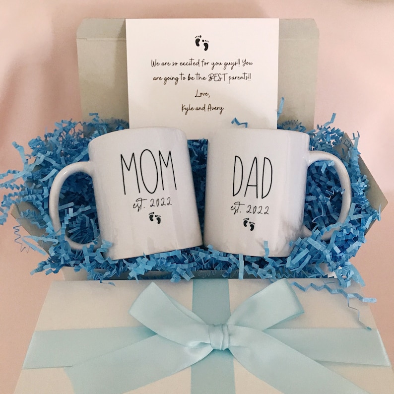 May include: A white gift box with a blue ribbon tied in a bow. Inside the box are two white mugs with black lettering. One mug says "MOM est. 2022" and the other says "DAD est. 2022".
