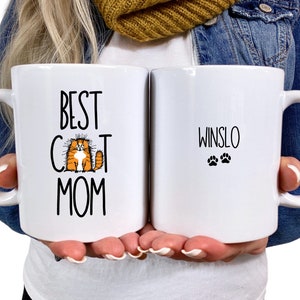 May include: Two white ceramic mugs. One mug says "Best Cat Mom" with a cartoon orange cat. The other mug says "Winslo" with two paw prints.