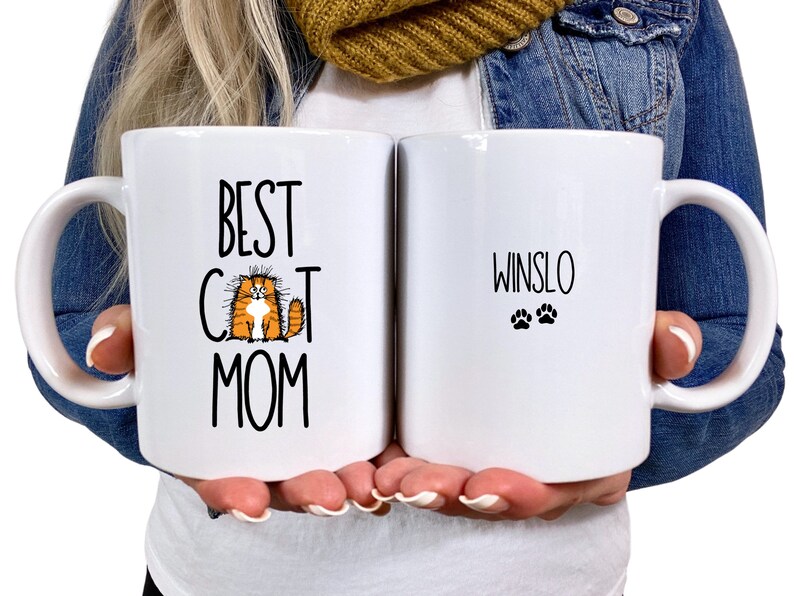 May include: Two white ceramic mugs. One mug says "Best Cat Mom" with a cartoon orange cat. The other mug says "Winslo" with two paw prints.