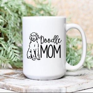 Op de afbeelding: Witte keramische koffiemok met een zwarte doodle-hond illustratie en de tekst "Doodle MOM" erop gedrukt.