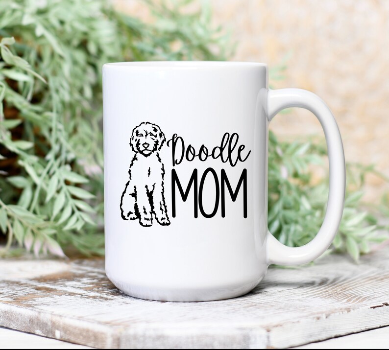 Op de afbeelding: Witte keramische koffiemok met een zwarte doodle-hond illustratie en de tekst "Doodle MOM" erop gedrukt.