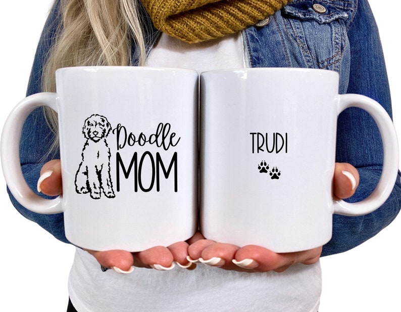 Op de afbeelding: Witte keramische koffiemok met een zwarte doodle-hond-illustratie en de tekst "Doodle Mom" aan de ene kant en de tekst "Trudi" met twee poot afdrukken aan de andere kant.