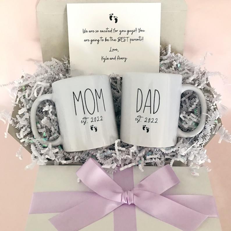 May include: A white gift box with a purple ribbon and two white mugs. The mugs have the words "MOM est. 2022" and "DAD est. 2022" printed on them. The box also includes a card with the message "We are so excited for you guys!! You are going to be the BEST parents!! Love, Kyle and Avery."