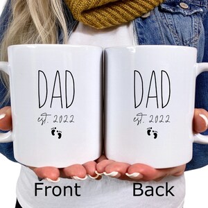 May include: Two white ceramic mugs with black text and a footprint design. The text on the mugs reads "DAD est. 2022".