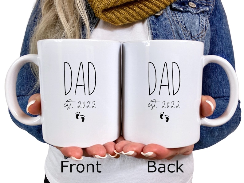 May include: Two white ceramic mugs with black text and a footprint design. The text on the mugs reads "DAD est. 2022".