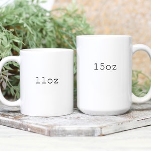 May include: Two white ceramic mugs of different sizes are displayed on a wooden surface. The smaller mug has "11oz" printed on it, while the larger mug has "15oz" printed on it. Green foliage is visible in the background.