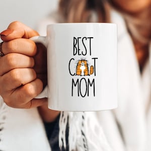 May include: White ceramic mug with black text that reads "Best Cat Mom" and a cartoon orange cat with black eyes.