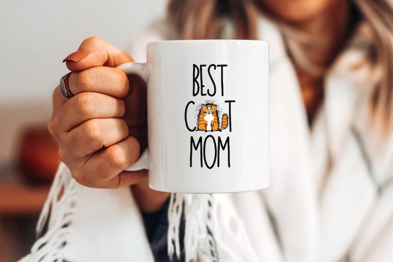 May include: White ceramic mug with black text that reads "Best Cat Mom" and a cartoon orange cat with black eyes.