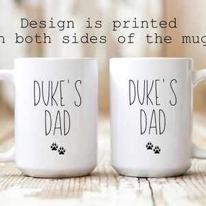 May include: Two white ceramic mugs with black text that reads "Duke's Dad" and two black paw prints on each mug.