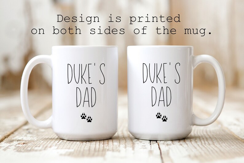 May include: Two white ceramic mugs with black text that reads "Duke's Dad" and two black paw prints on each mug.