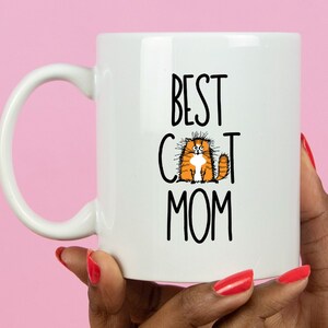 May include: White ceramic mug with a black and white graphic that says "Best Cat Mom" and a cartoon orange cat with white fur on its chest.