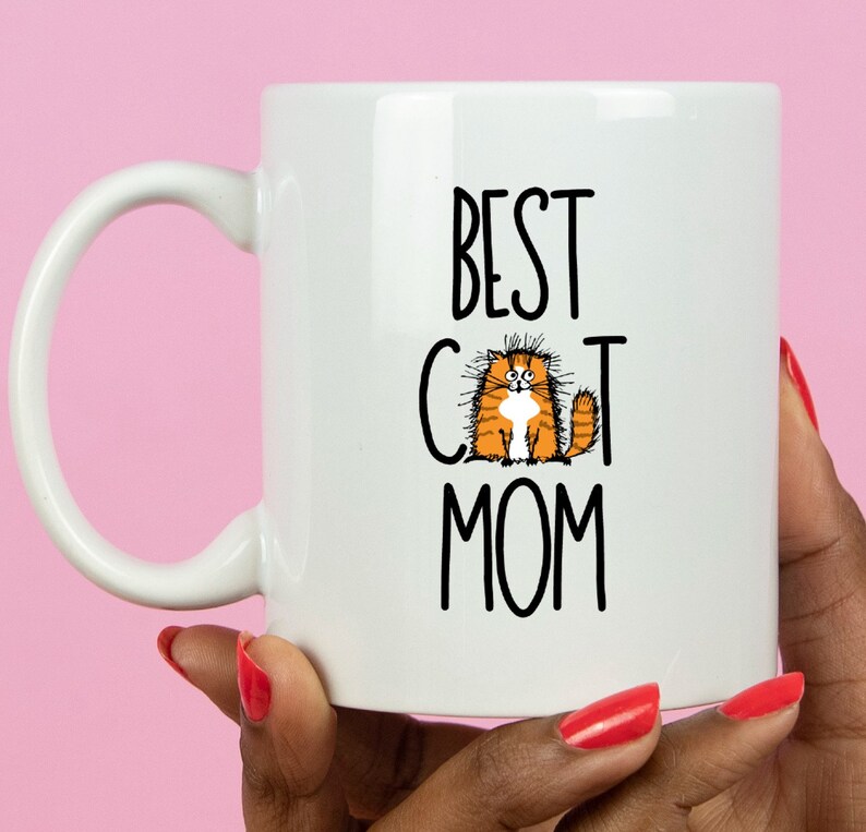May include: White ceramic mug with a black and white graphic that says "Best Cat Mom" and a cartoon orange cat with white fur on its chest.