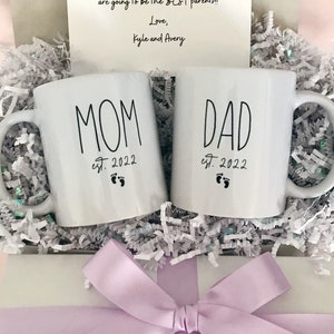 May include: Two white ceramic mugs with black lettering. The mug on the left says "MOM est. 2022" with a footprint symbol. The mug on the right says "DAD est. 2022" with a footprint symbol. The mugs are in a white box with light purple tissue paper and a light purple bow.