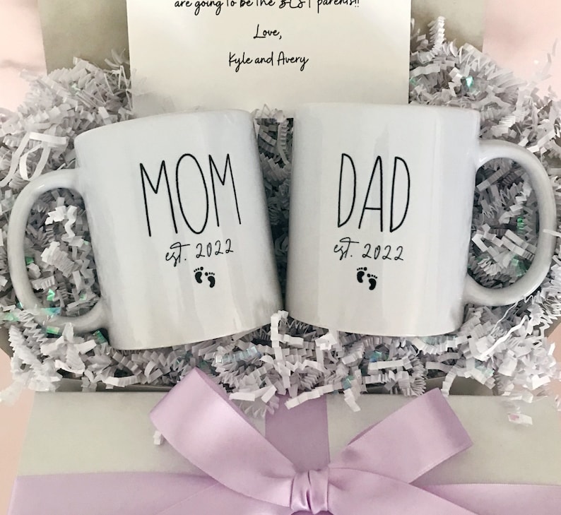 May include: Two white ceramic mugs with black lettering. The mug on the left says "MOM est. 2022" with a footprint symbol. The mug on the right says "DAD est. 2022" with a footprint symbol. The mugs are in a white box with light purple tissue paper and a light purple bow.