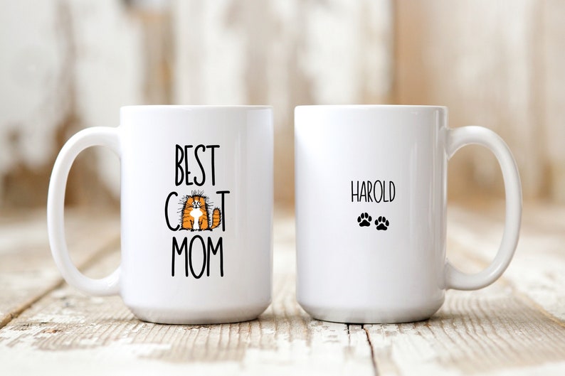 This white ceramic mug is available in 11 or 15 ounces. Black font is used. The words Best Cat Mom are on the front, along with an adorable orange cat in place of the letter A in cat. The back is personalized with the cats name, and tiny paw prints.