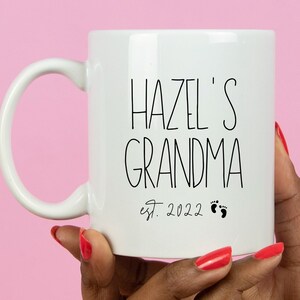 Op de afbeelding: Witte keramische mok met zwarte tekst die "Hazel's Grandma est. 2022" met twee kleine zwarte voetafdrukken.