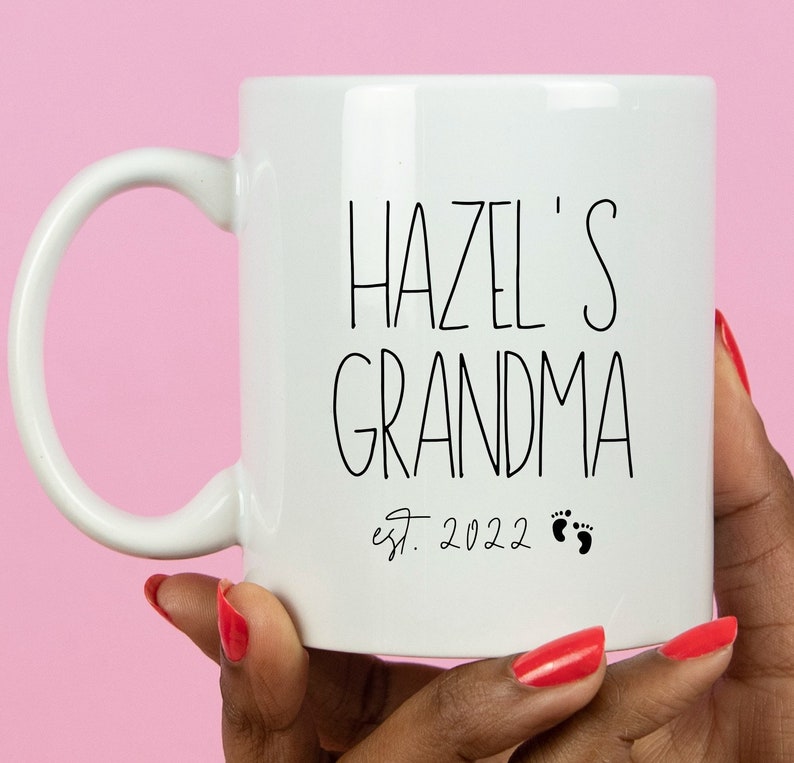Op de afbeelding: Witte keramische mok met zwarte tekst die "Hazel's Grandma est. 2022" met twee kleine zwarte voetafdrukken.