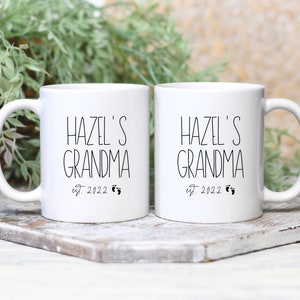Op de afbeelding: Twee witte keramische mokken met zwarte tekst die "Hazel's Grandma est. 2022" luidt en een kleine voetafdruk afbeelding.