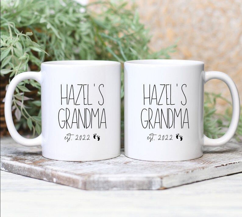 Op de afbeelding: Twee witte keramische mokken met zwarte tekst die "Hazel's Grandma est. 2022" luidt en een kleine voetafdruk afbeelding.