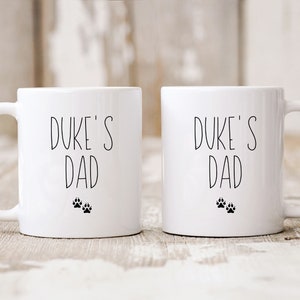 May include: Two white ceramic mugs with black text and paw prints. The text on both mugs reads "Duke's Dad".