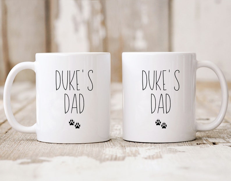 May include: Two white ceramic mugs with black text and paw prints. The text on both mugs reads "Duke's Dad".