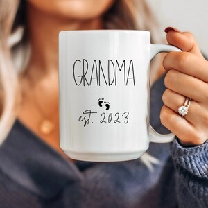 May include: White ceramic mug with the word "GRANDMA" in black script, accompanied by baby footprints and the year "est. 2023". The mug has a large handle and is held by a person.