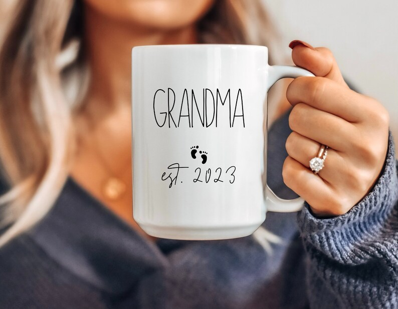 May include: White ceramic mug with the word "GRANDMA" in black script, accompanied by baby footprints and the year "est. 2023". The mug has a large handle and is held by a person.