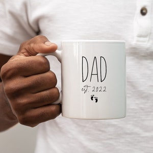 May include: White ceramic mug with black text that reads "DAD est. 2022" with two small footprints below the text.