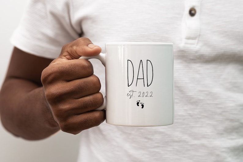 May include: White ceramic mug with black text that reads "DAD est. 2022" with two small footprints below the text.