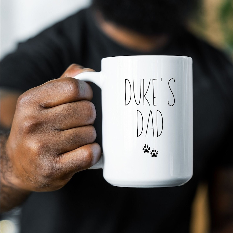 Man holding a white ceramic mug that reads Duke's Dad and includes small dog prints below the word Dad. The design is printed on both sides of the mug.