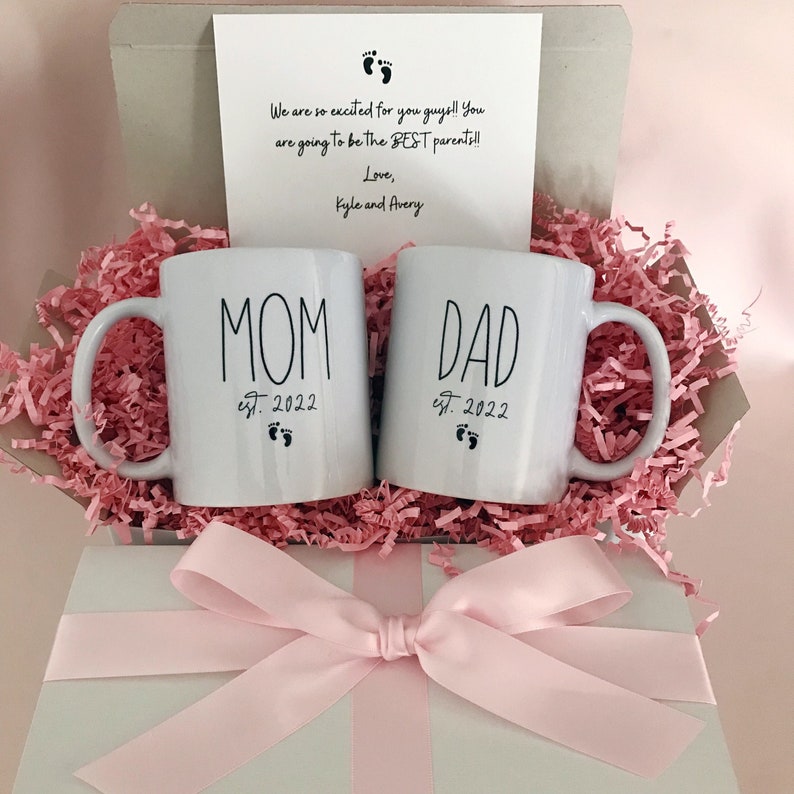 May include: A white gift box with pink tissue paper and a pink ribbon. The box contains two white mugs with black lettering. One mug says "MOM est. 2022" and the other says "DAD est. 2022".