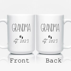 May include: Two white ceramic mugs with the word "GRANDMA" in black script, accompanied by baby footprints and the text "est. 2023." The front of one mug is labeled "Front," and the back of the other mug is labeled "Back."
