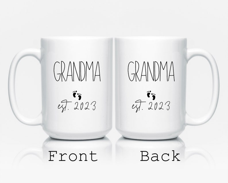 May include: Two white ceramic mugs with the word "GRANDMA" in black script, accompanied by baby footprints and the text "est. 2023." The front of one mug is labeled "Front," and the back of the other mug is labeled "Back."