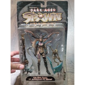 Könnte beinhalten: Todd McFarlane's Dark Ages Spawn Ultra Action Figures Blisterverpackung mit einer Kriegerfigur mit gehörntem Kopfschmuck, langem blondem Haar und dunkler Rüstung. Enthält eine skelettartige Kreatur, einen großen Stab und ein türkisfarbenes Sensen-Zubehör.