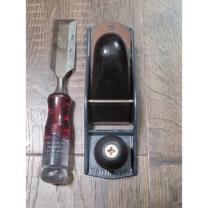 May include: A wood chisel with a translucent red handle and a metal blade alongside a block plane featuring a dark gray body, a black handle, and a black adjustment knob. The chisel handle displays text including "MADE IN USA" and "WARNING".