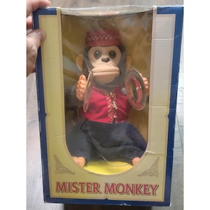 Puede incluir: Un juguete Mister Monkey en una caja rectangular. El mono tiene pelaje oscuro, cara color canela y viste un chaleco rojo con adornos dorados, un sombrero rojo y pantalones oscuros. Sostiene dos platillos de metal plateado en sus manos. Caja azul y amarilla.