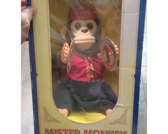 May include: A Mister Monkey toy in a rectangular box. The monkey has dark fur, a tan face, and wears a red vest with gold trim, a red hat, and dark pants. It holds two silver-colored metal cymbals in its hands. The box is blue and yellow.