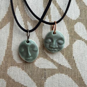 May include: Two ceramic pendants with serene face designs. Each pendant is a soft green color and hangs from a black cord. One pendant has a silver-colored bail, and the other has a copper-colored bail. The background has a neutral pattern.