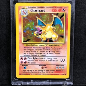 May include: A holographic Charizard Pokemon card from the 1999 Wizards set. The card is in a protective case. The card features a red and orange Charizard with blue wings and a yellow background. The card text reads "Charizard 120 HP" and "Fire Spin 100".