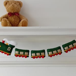 May include: A felt train garland featuring a green locomotive with white smoke and four green passenger cars with red wheels. The train is strung on a white cord decorated with small, colorful triangular felt pennants in teal, pink, and yellow.