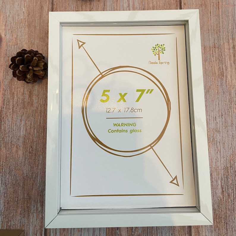 K&ouml;nnte beinhalten: Wei&szlig;er Bilderrahmen mit goldener Umrandung und einem goldenen Kreis mit dem Text "5 x 7" und "12,7 x 17,8 cm" darin. Der Text "WARNUNG Enth&auml;lt Glas" befindet sich unter dem Kreis. Der Text "Needle Spring" befindet sich in der oberen rechten Ecke des Rahmens.